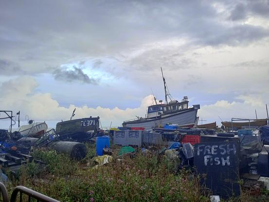 Fishing boats on a shoreline surrounded by pots, nets and things. A sign says Fresh Fish. Sky is all colours of grey with a clear ring of blue centre left.