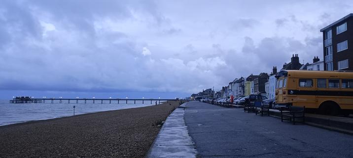 Nightfall at a beach promenade with a yellow bus in the carpark on right, a pier in sea on left, cloudy, all very grey