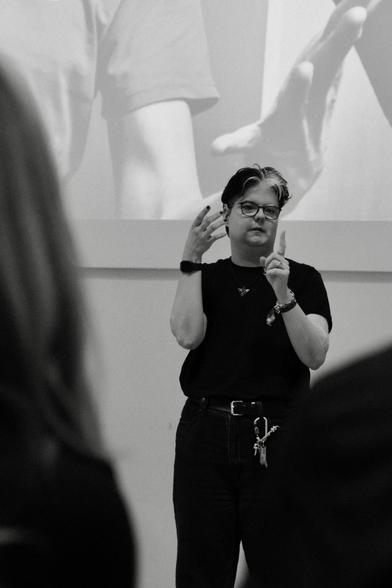 Dieses Bild zeigt mich (weiße Person) in einem Vorlesungssaal. Das Bild ist in Schwarz-Weiß aufgenommen, was den Fokus auf die Gestik und Mimik der Person legt.

Die Person trägt eine Brille und hat kurzes Haar mit einer markanten weißen Strähne an der Vorderseite. Person trägt ein schwarzes T-Shirt und dunkle Hosen. Auffällig ist der Schlüsselbund, der an der Gürtelschlaufe hängt.