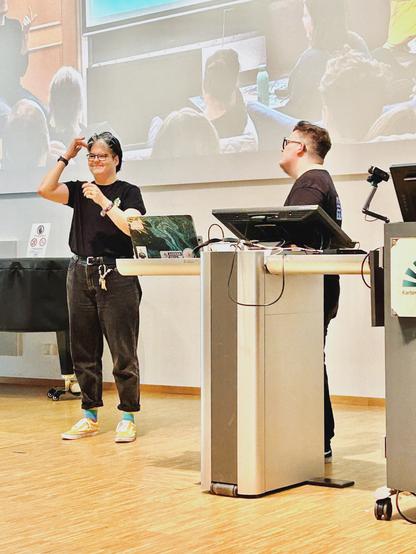 Dieses Bild zeigt zwei weiße Personen, die in einem Vorlesungssaal stehen. Eine Person links (ich) gebärdet mit der Hand in der Nähe des Gesichts, während die andere Person rechts steht und mich anschaut. Beide tragen schwarze Kleidung.
Im Vordergrund sieht man ein Rednerpult mit technischen Geräten wie Computern und Mikrofonen. Es sind Sticker auf dem Laptop zu erkennen. Die Bühne ist sauber und professionell eingerichtet.
Im Hintergrund wird eine Projektion des Geschehens auf einer Leinwand angezeigt, wo ein Foto vom Sensibilisierungsworkshop zu Gehörlosigkeit eingeblendet wird.