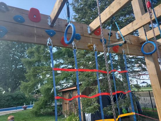 Different colour climbing holds in a zig zag across a 2x8 beam of wood on a bigger structure. A colourful cargo net hangs below. A grass back yard is in the background.