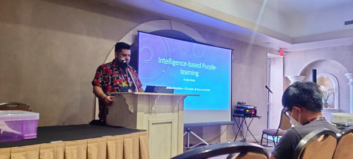 A speaker at a conference presents a talk on ‘Intelligence-based Purple-teaming’ while standing at a podium. A slide with the presenter’s name, Carlos Gonçalves, is visible on a screen behind them. The room has a professional setting with chairs and a sound system.