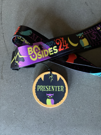 A close-up image of a conference badge with a lanyard. The badge has a circular design with the word ‘PRESENTER’ printed in bold green text against a dark background. The lanyard attached to the badge reads ‘BSides Las Vegas 24’ in colorful letters, decorated with various patterns.