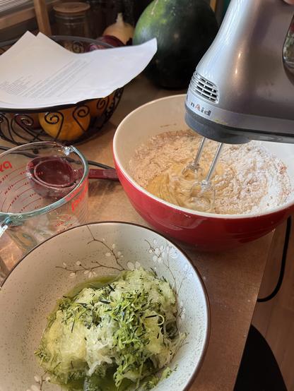 Photo showing a bowl of shredded zucchini in the foreground, and an active hand mixer in the background, mixing a wet dough mixture. Off to the side is a Pyrex measuring cup, some dirty plastic measuring cups, and a printed recipe.