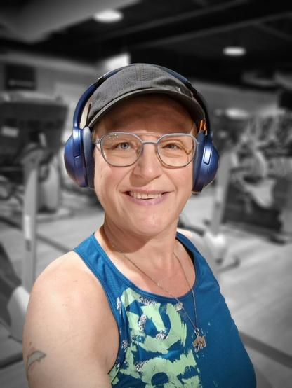 Selfie of me in the gym, riding the seated bike. Smiling at the camera. Used a filter to have me in color and the background is black and white. Wearing a blue top with green accents, black cap, blue headphones and blue-rimmed glasses. In the background are several cardio machines.