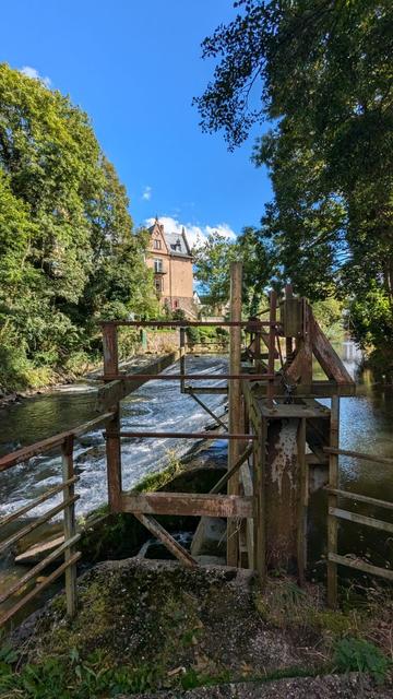 Ein kleiner Fluss, die Urft, überfließt ein Wehr. Im Vordergrund die verrostete Wehranlage. Links und rechts des Flusses grüne Bäume, darüber blauer Himmel.