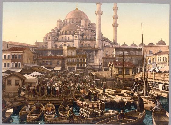 This image is a postcard depicting the bustling life of Istanbul's Eminönü bazaar and Yeni Cami (New Mosque) around 1890-1900. The scene is vibrant with people engaged in various activities, creating an atmosphere of liveliness and commerce. The buildings have distinct Ottoman architectural features, such as the domes and minarets that are characteristic of this style. The color palette consists mainly of earth tones, providing a sense of warmth and history to the scene.