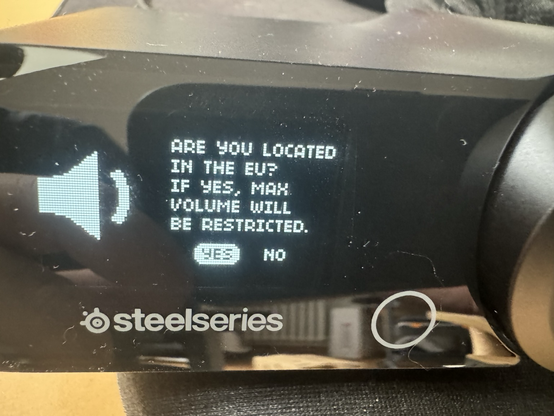 Photo of a black plastic appliance with a volume knob to the left and a small LCD pixel display. Text next to a loudspeaker icon reads: "Are xou located in the EU? If yes max. Volume will be restricted." Below that there's a YES/NO prompt.
