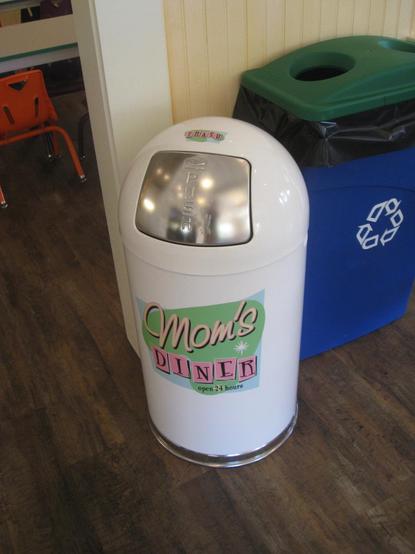 A white trash can labeled "Mom's Diner" with a circular silver push-top lid, positioned next to a blue recycling bin. The setting has wooden flooring and hints of a diner ambiance.
