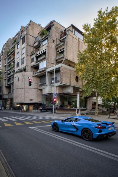 Ein moderner Wohnbau aus Beton und Backsteinen. Auf der Straße davor ein blauer Sportwagen.