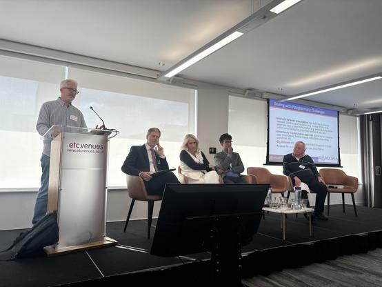 A panel discussion taking place in a conference room. A speaker - Mike - is at a podium addressing an audience, while four panelists sit on stage with a presentation about polypharmacy challenges displayed on a screen behind them.