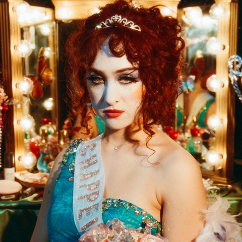 Album cover for The Rise and Fall of a Midwest Princess by Chappell Roan. Image shows Roan wearing a blue dress with a sash with curly red hair and a tiara looking intensely with a brightly lit vanity behind her.