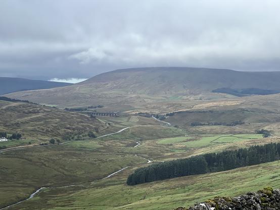 A scenic landscape featuring rolling hills and valleys under a cloudy sky. A river winds through the foreground, and a viaduct is visible in the middle ground with fields and forests in the background.