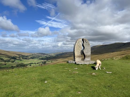 A scenic landscape featuring a stone sculpture set on a grassy hill. The sculpture has a curved, split design. A sheep is grazing nearby, with rolling hills and a blue sky with scattered clouds in the background.