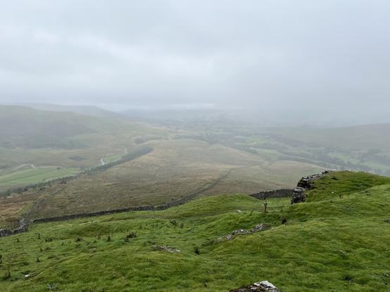 A misty landscape featuring rolling green hills, valleys, and winding roads. The sky is overcast, creating a gloomy atmosphere. Stone walls are visible in the foreground, and distant hills fade into the fog.