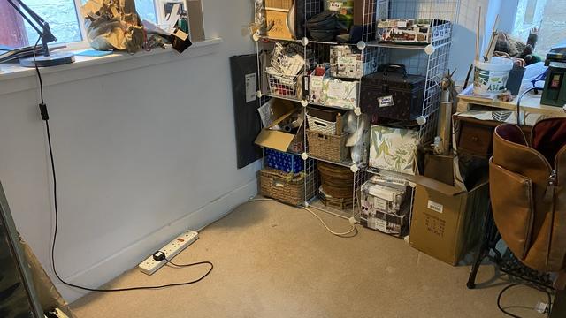 A corner of a room featuring a stack of storage baskets and boxes, a desk with a wooden finish, and a lamp. Various craft supplies and materials are visible on the shelves, along with some stacked boxes and a power strip on the floor. The carpet is a soft honey colour and has been hoovered clean!