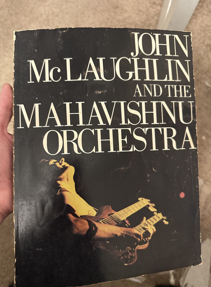 JOHN MCLAUGHLIN AND THE MAHAVISHNU ORCHESTRA. Man with double neck guitar strung in 12 and 6 strings plays and looks up in a dark stage lit in yellow.