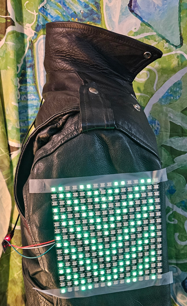 Side of a leather biker's jacket. An LED matrix panel is taped to the side. Wires to a microcontroller can be seen hanging off to the left. The LEDs show a cascading chevron pattern.