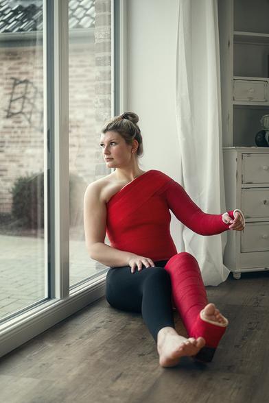 Eine junge Freu sitzt mit einem wehmütigen Blick nach draußen vor einem bodentiefen Fenster. Ihr ganzes linkes Bein, der linke Arm, Schulter und Oberkörper stecken in roten Kunststoffgipsen, am anderen Bein trägt sie eine schwarze Leggings.