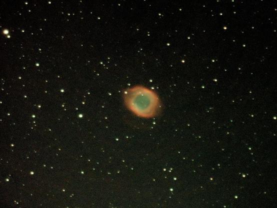 A sparse starfield frames the startling cerulean eye of the Helix Nebula, an oval shaped planetary nebula with a blue green iris like centre. It is rimmed by orange reddish annular surround.