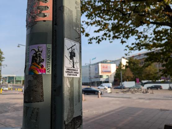 Two stickers on a utility pole in Helsinki. In the background, the Kiasma Museum of Contemporary Art is visible with a banner for the Simon fujiwara exhibition. 

The sticker on the left depicts Goatman in a three quarter profile with a rainbow cloak. The inscription "Always Antifascist"  is above and below. 

On the right is a Greenbelt Utility Pole Fan Club sticker showing   the World Famous Greenbelt Utility Pole in all of its splendor.