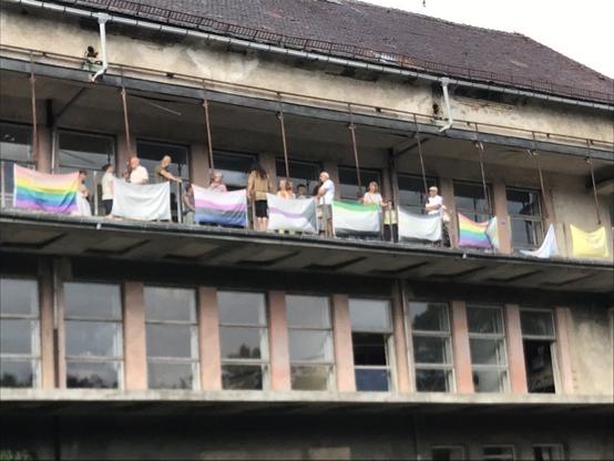Häuserfront mit zwei Reihen Balkon und Fenstern, auf dem oberen steht eine Gruppe Menschen. Die Front ist mit verschiedenen Pride Flags versehen. Das Gebäude sieht alt und etwas verfallen aus.