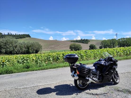A big black touring bike parked on the side of a country road. The typical tuscany panorama with green hills and a clear blue sky in the background. Some very low and little clouds are placed along the skyline.