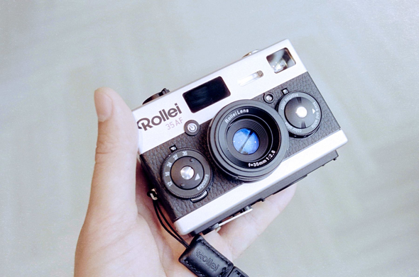 A photo of a small boxy camera in hand on a light, blurry background. Lens faces the camera