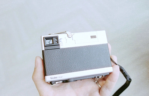 A photo of a small boxy camera in hand on a light, blurry background. Back plate faces the camera