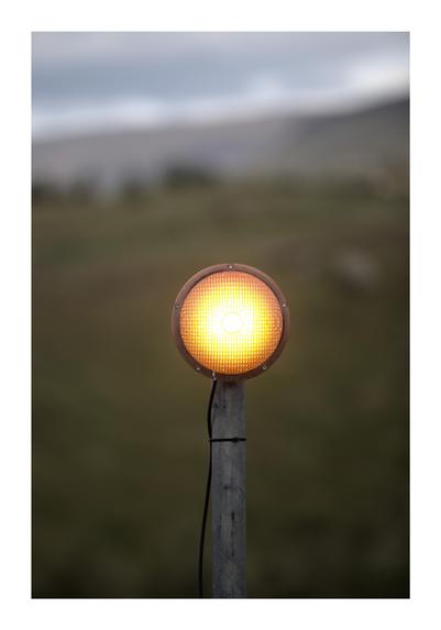 A close-up image of a single yellow-orange traffic or signal light mounted on a pole. The light is glowing against a blurred, natural landscape background with soft green tones. The simplicity of the scene creates a contrast between the man-made light and the natural setting.