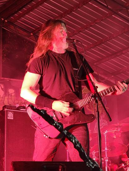 Mark Kloeppel of Misery Index. He is on stage performing guitar and backing vocals. Red light is shining everywhere.