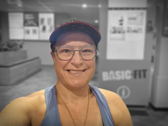 Selfie of a person, sitting in the relaxing area of the gym. Smiling at the camera. Used a filter to have me in color and the background is black and white. Wearing a blue top, black cap and blue-rimmed glasses.