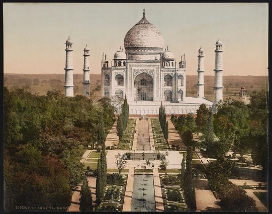 The image depicts the majestic Taj Mahal in Agra, India. This historical monument is known for its large white marble dome and four slender minarets that reach into the sky. The perspective of the photograph shows a front view of the Taj Mahal, surrounded by lush green trees. In the foreground, there are paths leading to the entrance gate of the complex, inviting visitors to explore this world-renowned landmark.