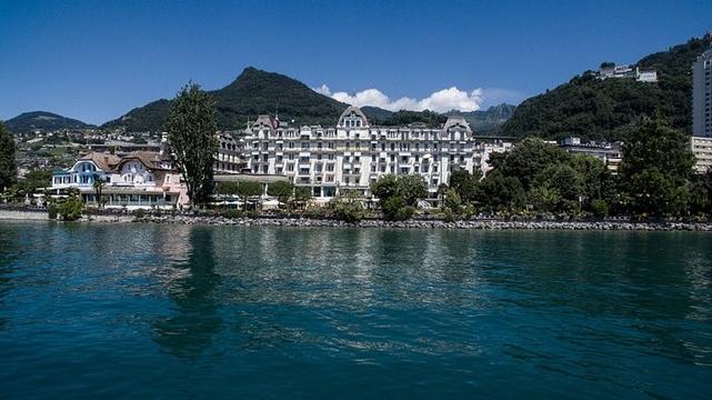 Photo of a majestic old hotel on the waterfront with great mountains and a blue sky in the background.