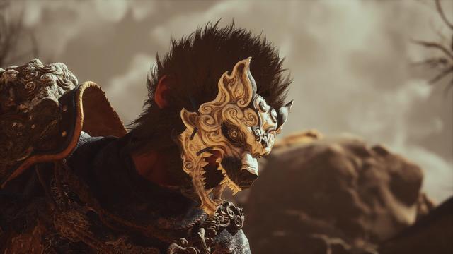 The image shows a close-up of the Black Myth WuKong character with an intricately carved mask that resembles a terrifying animal face. The mask has sharp teeth and a detailed, ornate design. The warrior wears richly decorated armor and has disheveled, dark hair. The background is blurred but shows a rocky landscape under a cloudy sky, creating a dramatic mood.