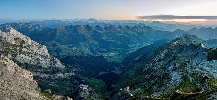 Das Bild zeigt eine weitläufige Panoramalandschaft in den Alpen. Im Vordergrund sind imposante, zerklüftete Felsformationen zu sehen, die steil ins Tal abfallen. Das Tal selbst ist üppig grün und durchzogen von kleinen Dörfern und Wiesen. In der Ferne erstreckt sich eine lange Bergkette mit vielen Gipfeln, die sich im Dunst des Horizonts verlieren. Der Himmel ist klar mit sanften Pastellfarben am Horizont, die auf den beginnenden Sonnenaufgang oder Sonnenuntergang hindeuten. Insgesamt strahlt die Szene Ruhe und Erhabenheit aus, während die Berge und das Tal im sanften Licht erstrahlen.