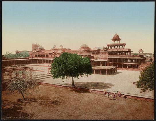 The image depicts a historic building complex, likely in India. It appears to be an old photograph, characterized by the sepia tones and faded appearance typical of such historical images. The architecture suggests that it could be a palace or mahal from ancient times, with multiple levels of structures and intricate details on pillars and walls. There is also a tree visible in front of the building, providing some greenery to the scene. The style of the image and the attire of any figures present would suggest it was taken around the turn of the 20th century.