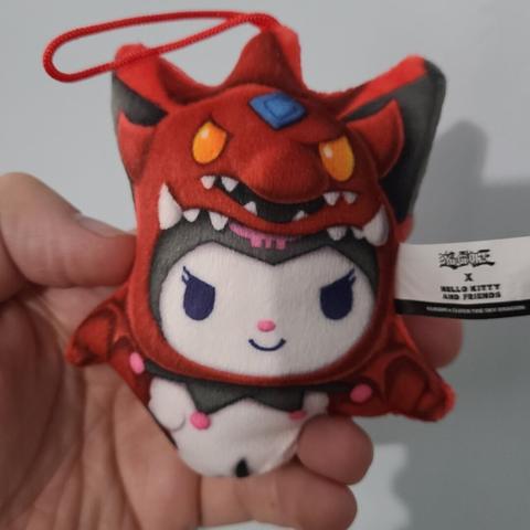 Photograph of an adult white person's hand holding a small plush figure of Kuromi (white bunny with angry eyes and a black hood) dressed as the Silfer the Sky Dragon from Yu Gi Oh (red dragon with a blue gemstone in the center of their head and yellow eyes).