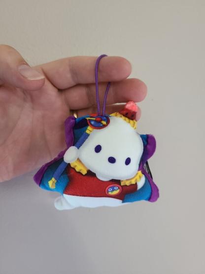 Photograph of an adult white person's hand holding a small plush figure of Pochacco (white puppy with dark colored ears) dressed as the Time Wizard from Yu Gi Oh (wearing a red suit with a blue / yellow button with eyes on it on the chest, a long cape, red hat, while holding a wizard's staff).