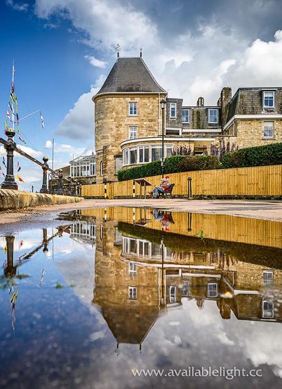 A large puddle reflecting a turreted building. A couple sitting on a bench in the distance, the gentleman wearing a red beret hat. The sky is blue with fluffy white clouds. The building sits on the seafront but the sea is out of shot behind railings and bunting.