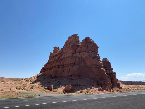 A stone sentinel stands in the open desert north of Hanksville, UT. Layers of sandstone eroded by the wind are visible on this shadowed side. Asphalt pavement (Hwy 12) is in the foreground, brilliant blue skies overhead. Was just so impressed at the size of this, which was once a much bigger mountain, that I had to pull over and take a picture.