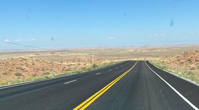 A ribbon of asphalt highway extends towards the horizon. The asphalt is marked with double yellow line in the center (no passing zone) and single white lines at the edges, marking the shoulder. Look closely and you'll see it turn and continue till it disappears into the brownish / greenish desert landscape full of sagebrush and remnants of eroded rock formations. Small utility poles follow the road carrying  barely perceptible wires. Distant hills come into view through the haze. Clear, light blue skies overhead.