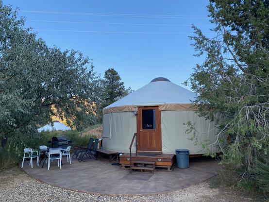 Photo shows a Yurt (circular tent like structure) topped by a dome-like skylight. A sturdy wooden front door and wooden steps welcomes one in. A concrete patio extends around the sides. To the left are 4 white chairs and table, a gas grill and a couple reclining chairs which make stargazing comfortable. Set among desert trees, the next yurt is visible in the distance.  Usual brilliant blue skies overhead.  Check out https://escalanteyurts.com/  for a highly recommended glamping experience.