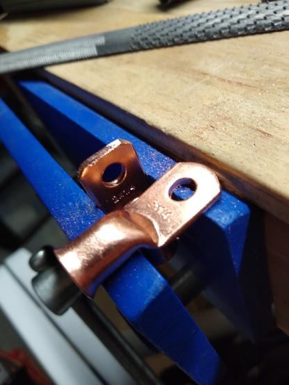 A copper wire lug clamped into a table vise. The lug has been filed down at the top edge to it's flat. A new lug sits in front of it for comparison.