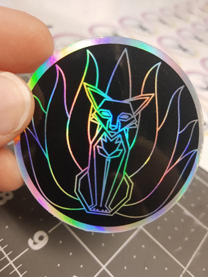 a circular sticker on holographic silver vinyl printed with black ink. Through the gaps in the black ink the silver makes the Arcane Whiskers logo of a kitsune (nine tailed fox)