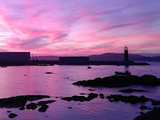 Photo of a sunset over the sea. The sky is sea and so it is the sea because it reflects the light from the sky. In the background there is a breakwater with a small lighthouse at the end. In the foreground there are some rocks in the sea but it's too dark to see anything else than the shape of those rocks.