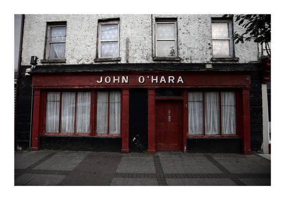 An old, weathered building with a faded red facade features the name "John O'Hara" in white letters above the door. The windows are covered with lace curtains, and the exterior shows signs of age, with peeling paint and worn walls. The building has a quaint, rustic feel, evoking a sense of history and nostalgia.