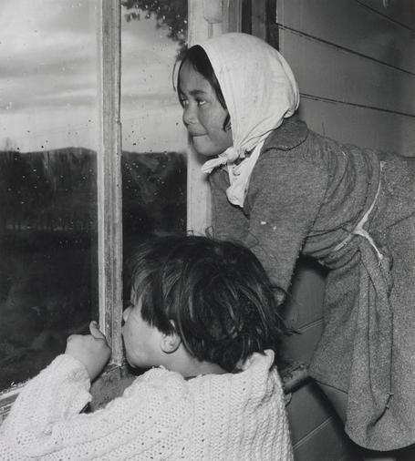 The image features two individuals looking out a window. On the left, there's an adult who appears to be wearing a headscarf and a patterned dress, while on the right, there's a child standing in front of the window. The window has raindrops on it, suggesting that it may have been raining recently. There's no clear text or other distinctive features visible in the image.