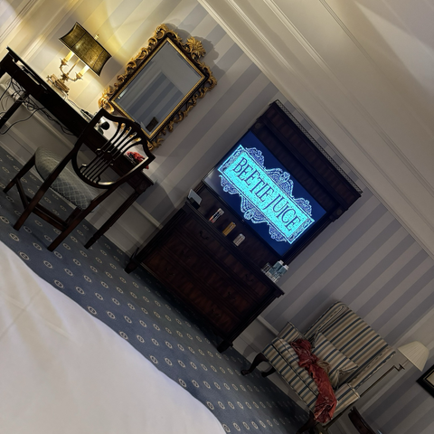 Hotelroom with a desk on the left, a tv-screen in the middle with Beeatlejuice on and a comfy chair on the right.