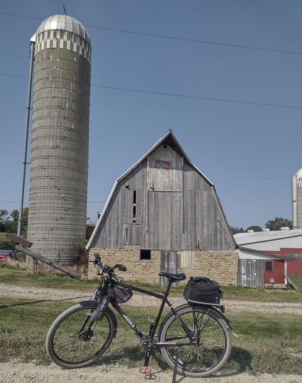 A black bicycle posed on the edge of a gravel road. A large old barn with dhrystone walls for the foundation and first floor, and the rest covered in vertically aligned planks is behind. At the peak of the barn, a worn painted Norwegian flag can just be made out in the shadows. To the side of the barn is a large, old, narrow silo many stores tall with a still bright metal roof. A clear blue sky is above the entire scene.